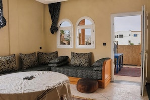 A cozy room with a sofa, a round table, and a wooden bench. There are two windows with arched tops and a door leading outside. - Seaview Apartment, Balcony, Great Location (Mirleft)
