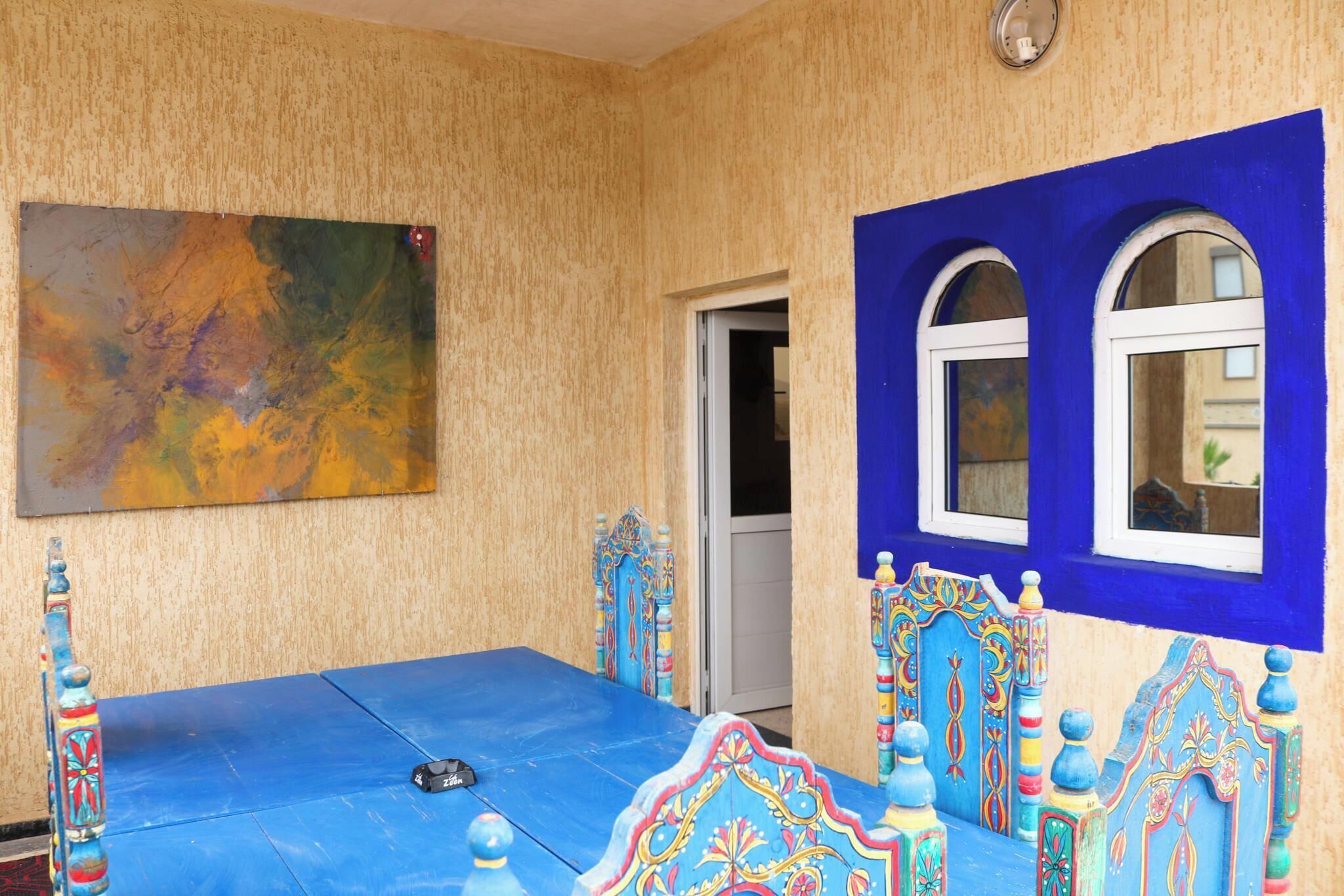 Een kamer met een blauw bed, een kleurrijk schilderij en een deur naar een andere kamer.