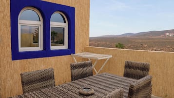 Un balcon agrémenté de meubles en osier, d’une petite table et de deux fenêtres en arc surmontées d’un cadre bleu.