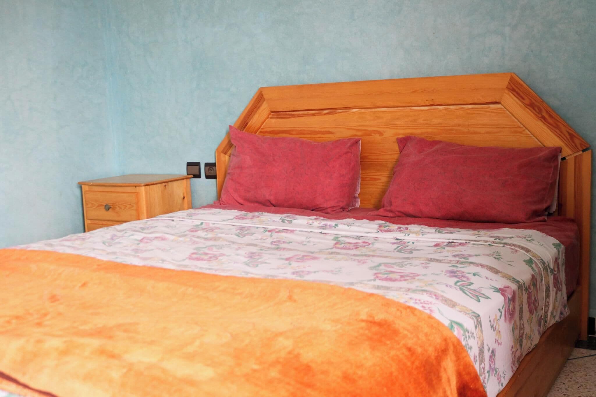 A bedroom with a wooden headboard, two pillows, a nightstand, and a floral bedspread.