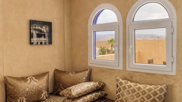 A room with a beige wall, two arched windows, a bed with patterned pillows, and a framed picture.