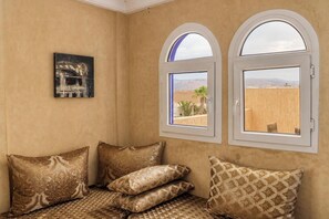A room with a beige wall, two arched windows, a bed with patterned pillows, and a framed picture.