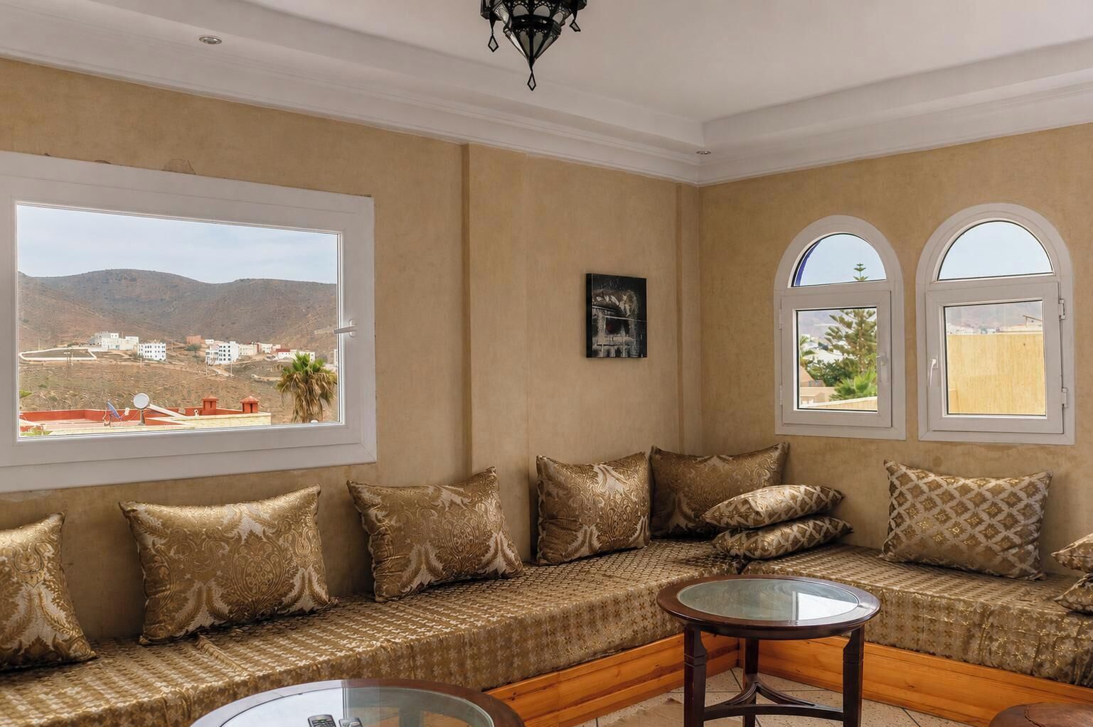 A room with a sofa set, a round table, and two windows offering a view of a landscape with buildings and hills.