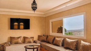 A room with a sofa, a small round table, a large window with a view, and a wall-mounted TV.