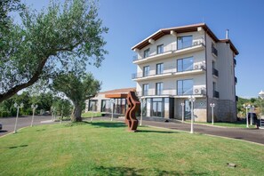 Un edificio de varios pisos con balcones, una entrada central y un área verde con una escultura.