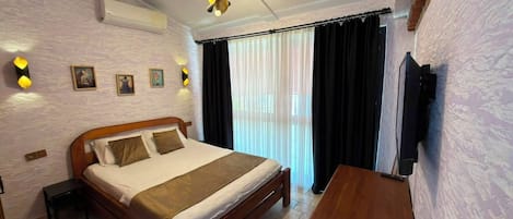 A hotel room with a bed, a bedside table, a flat-screen TV mounted on the wall, and a window with curtains.
