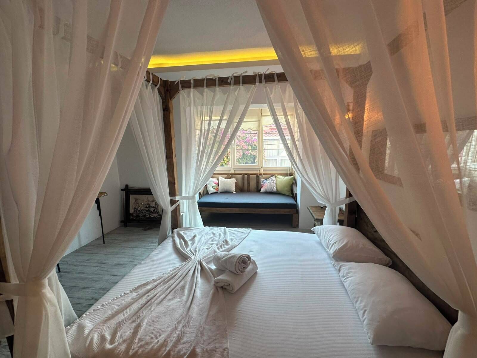 A bedroom with a canopy bed, a bench with cushions, and a view of the outside through a window.