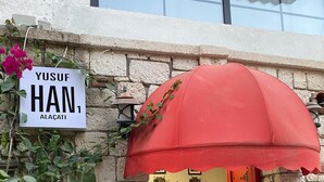 Entrada de um prédio de pedra, com um guarda-chuva vermelho, uma placa com a inscrição 'Yusuf Han Alacati' e um banco no interior.