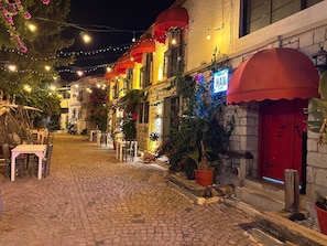 A cobblestone street lined with buildings, some with red awnings and outdoor seating. - Yusuf Han Alacati  (Cesme)
