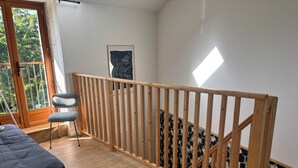 Ein Zimmer mit einer Holztreppe, einem Bett, einem Sessel und einem Fenster mit Blick ins Grüne.