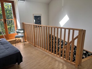 Ein Zimmer mit einer Holztreppe, einem Bett, einem Sessel und einem Fenster mit Blick ins Grüne.