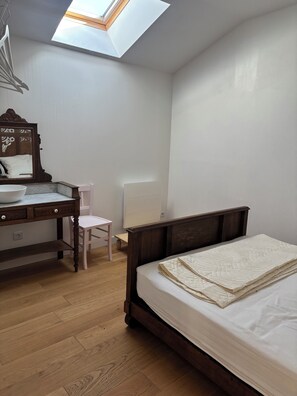 Une chambre avec un puits de lumière, un bureau en bois avec un miroir, une chaise blanche et un lit recouvert d’une couverture matelassée.