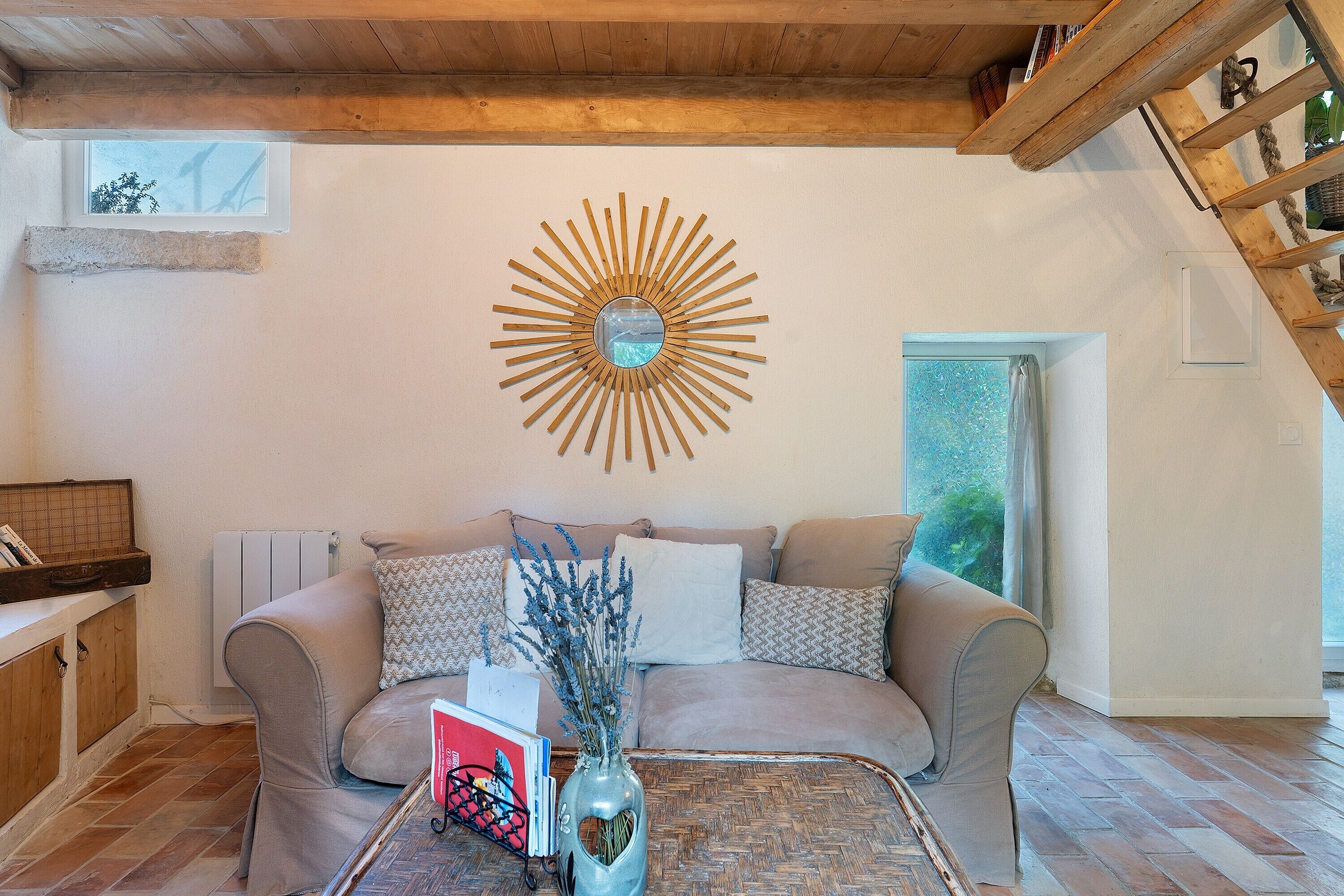 Un salon chaleureux avec un plafond en bois, une décoration murale en forme de soleil, un canapé agrémenté de coussins à motifs, une table basse sur laquelle est posé un vase de fleurs, et une vue sur la verdure à l’extérieur.
