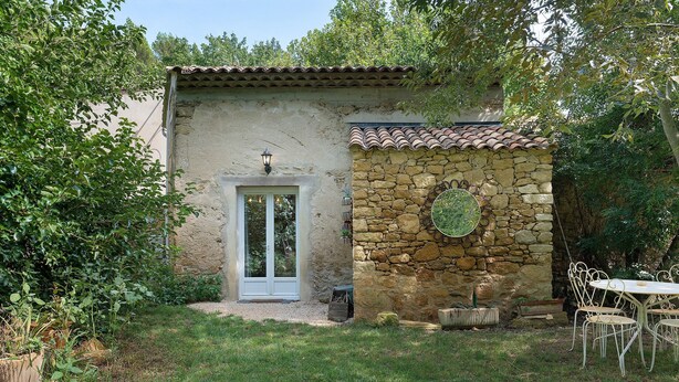 A rustic stone building with a tiled roof, a white door, and a small garden area with a table and chairs. - Holiday Home 'Maison En Pierre Chevaux' with Wi-Fi (Puyvert)
