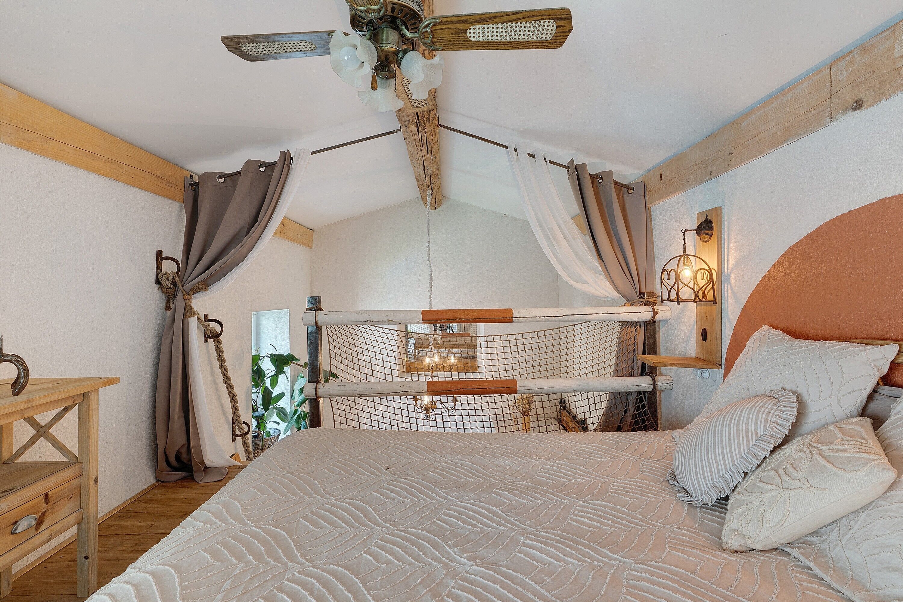 Une chambre à coucher avec un lit, des rideaux, une tête de lit en bois, une table de chevet et une lampe fixée au mur.