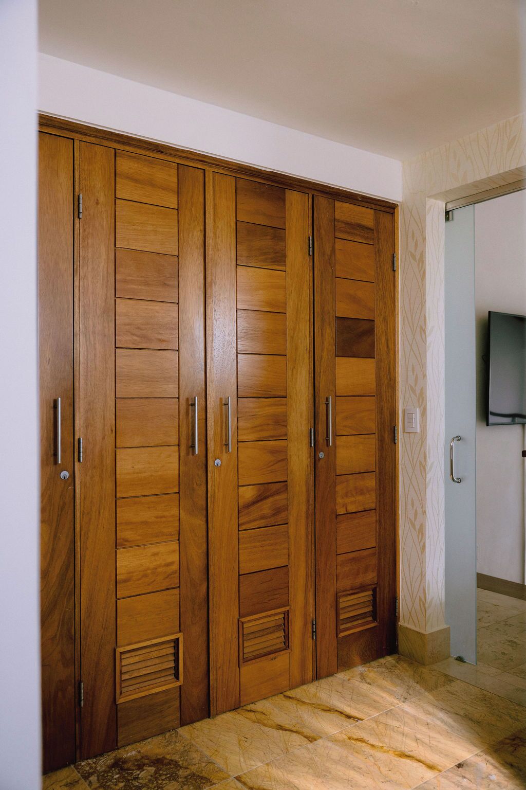 A wooden wardrobe with multiple doors and handles, a tiled floor, and a television mounted on the wall.