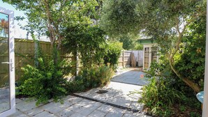 A paved patio area with a wooden fence, surrounded by lush greenery and trees. - Merrilees (Yarmouth)
