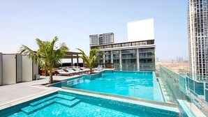 Ein Dachpool mit Liegestühlen und Palmen, umgeben von modernen Gebäuden.