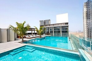 Ein Dachpool mit Liegestühlen und Palmen, umgeben von modernen Gebäuden.