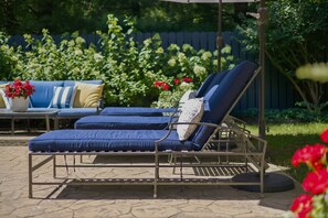 Une chaise longue avec une structure en métal et des coussins bleus, placée sur une terrasse en pierre, avec un jardin en arrière-plan.