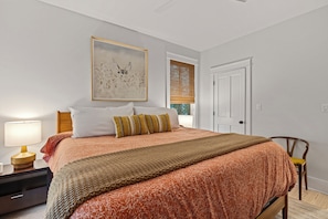 Une chambre à coucher avec un lit, une table de chevet avec une lampe, une chaise, une porte et une fenêtre avec des stores.