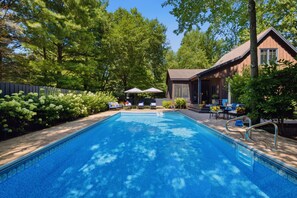 Une piscine entourée d’arbres et d’un bâtiment en bois, avec des chaises longues et des parasols à proximité.
