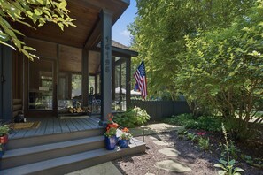 A covered porch with a wooden deck, a flagpole with an American flag, and a garden with stepping stones. - (2027 OPEN!) Gorgeous Union Pier Home With Private Pool! (Union Pier)
