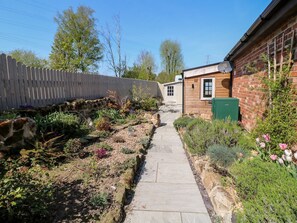 Ein Gartenweg führt zu einem Holgebäude mit einer grünen Tür, umgeben von Pflanzen und einer Steinmauer.