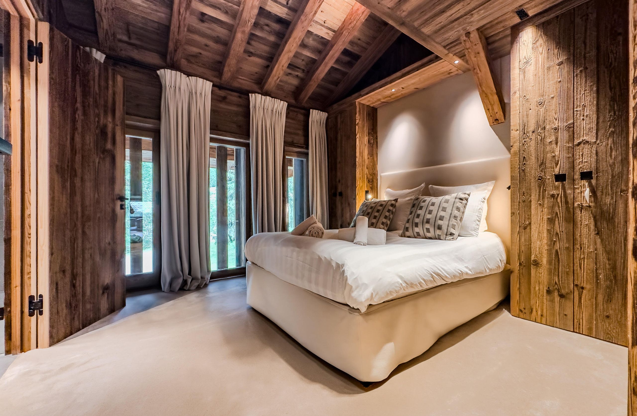 A bedroom with a large bed, wooden ceiling beams, and a view of greenery outside.