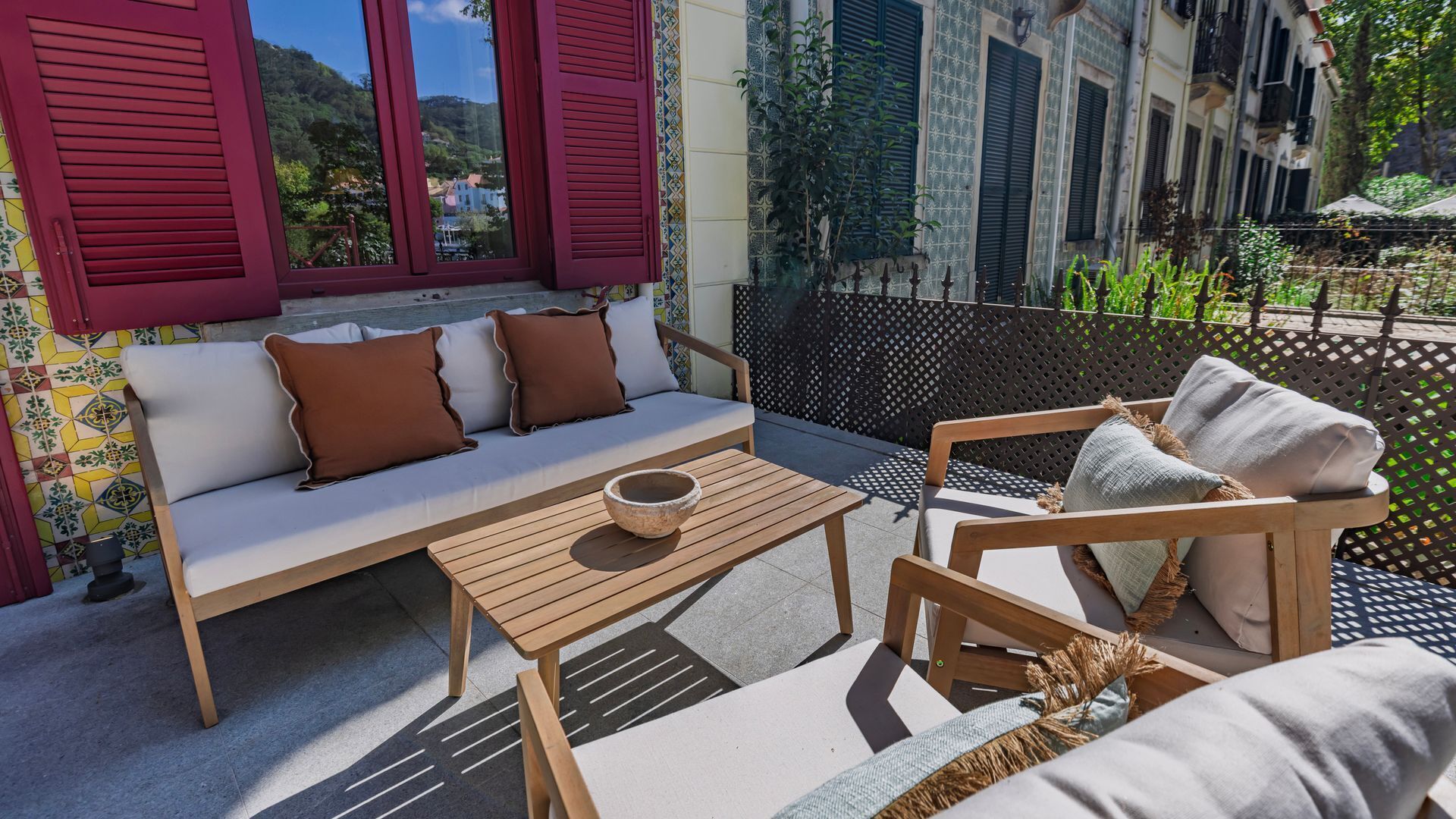 A sunlit outdoor seating area with a sofa, a wooden table, and a chair, featuring a view of a building with patterned tiles and shutters.