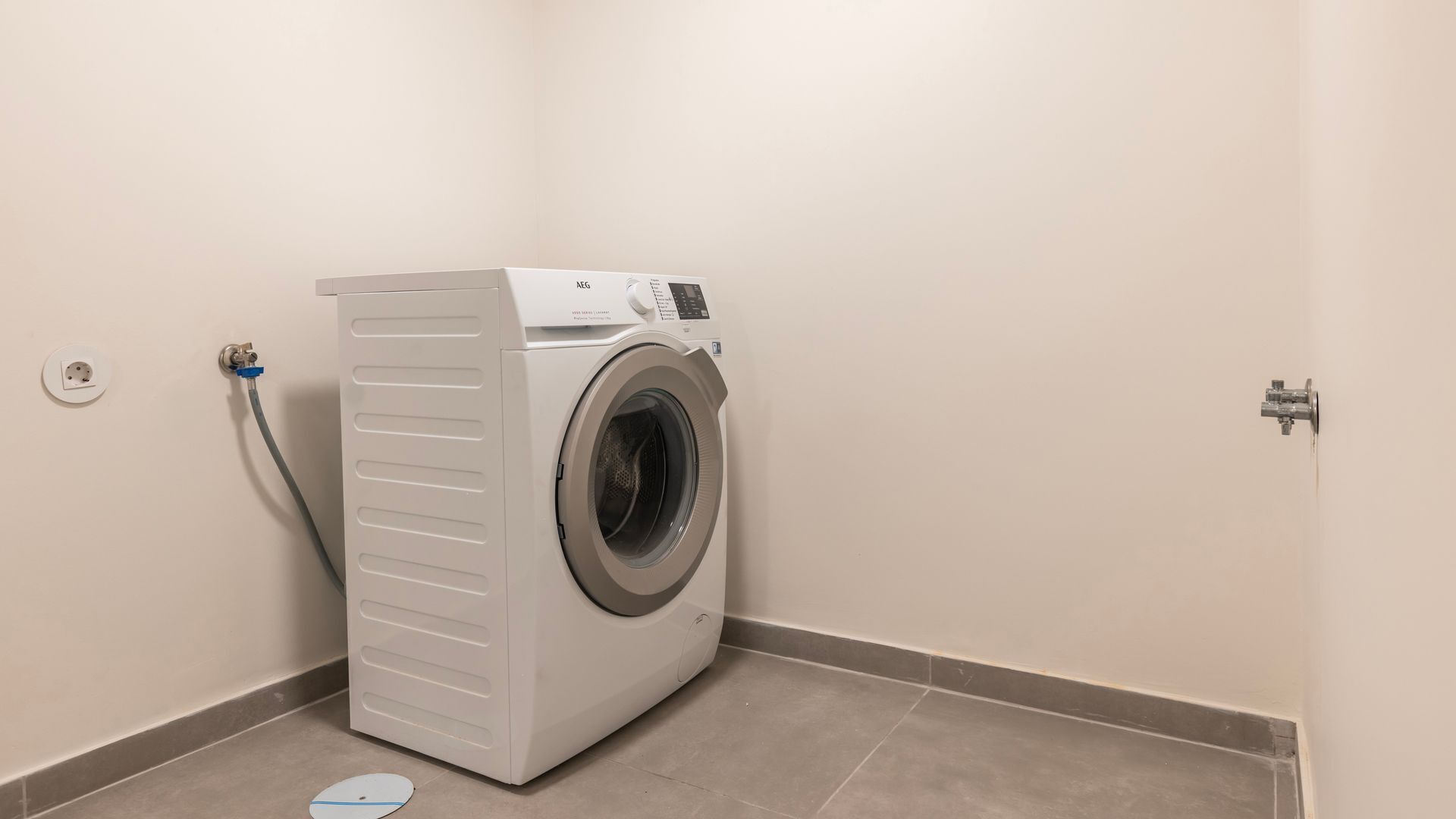 A compact, white front-loading washing machine in a small utility room with a tiled floor and a single electrical outlet.