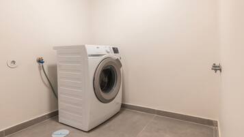 A compact, white front-loading washing machine in a small utility room with a tiled floor and a single electrical outlet.