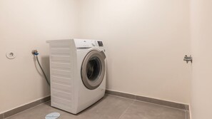 Una lavadora compacta de carga frontal, blanca, en un pequeño cuarto de lavado con piso de baldosas y un solo enchufe eléctrico.