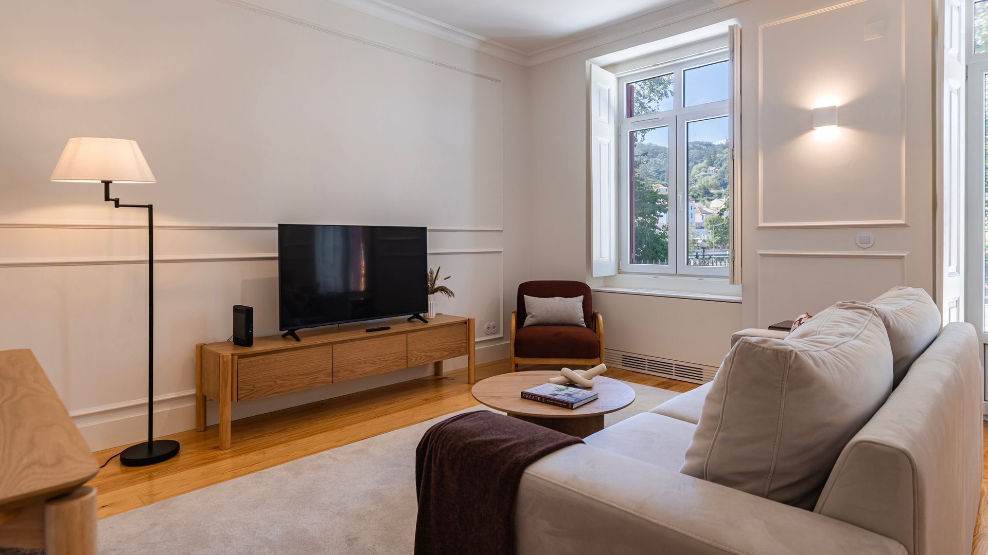 A living room with a flat-screen TV on a wooden console, a brown armchair, a beige sofa, and a lamp.