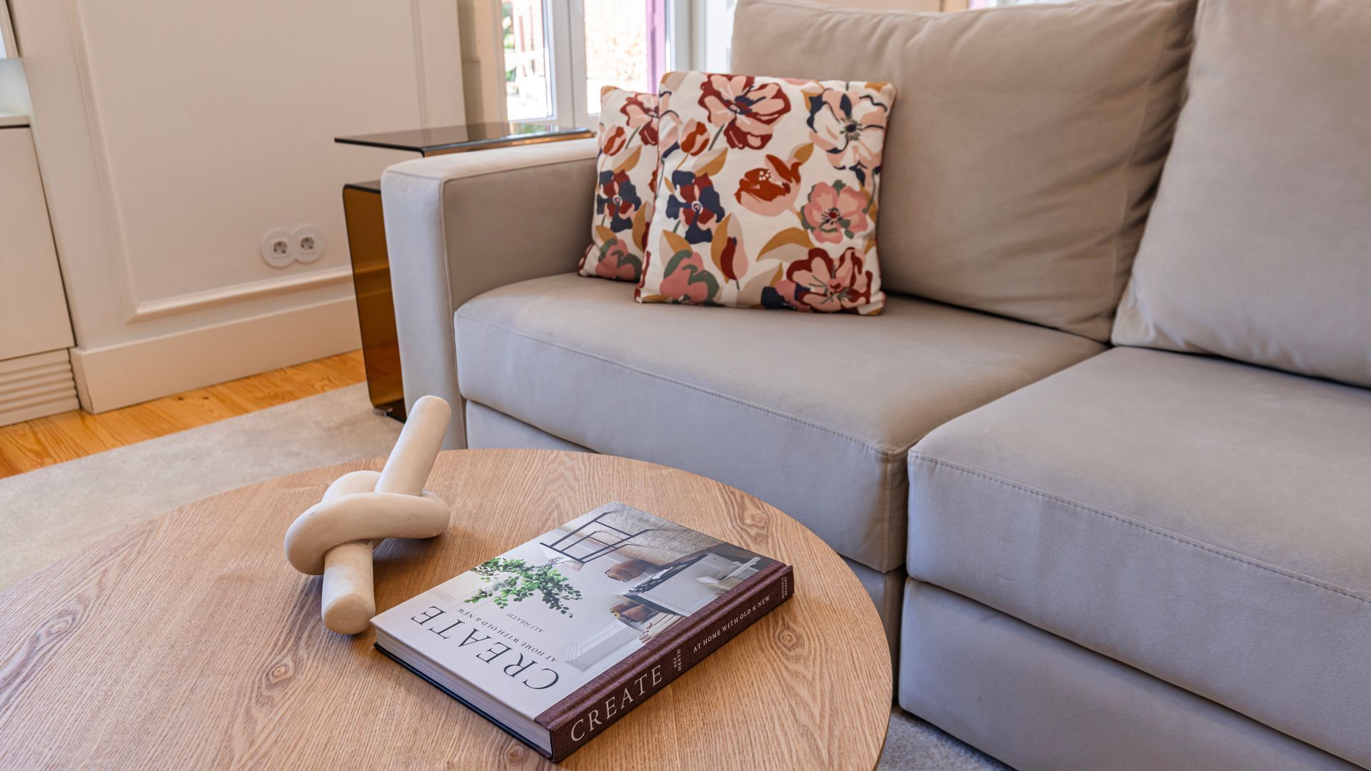 A modern living room with a grey sofa, a round wooden coffee table, and a book titled 'CREATE AT HOME WITH OLD & NEW'.
