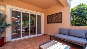 Eine sonnendurchflutete Terrasse mit einer Glastür, einem grauen Sofa, einer Holzbank und einem Blumentopf.