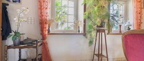 Une pièce avec une chaise en bois, une table ornée d’un vase de fleurs et une fenêtre avec des rideaux.