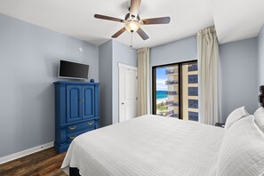 Ein Schlafzimmer mit einem Bett, einem Fernseher auf einer Kommode, einem Deckenventilator und Blick auf ein Gebäude mit Balkonen.