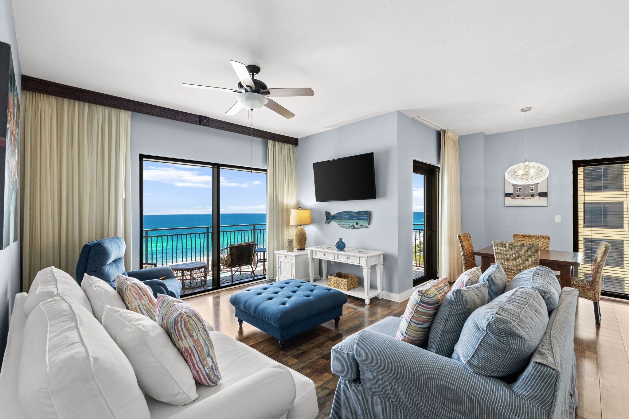 A spacious living room with a view of the ocean, a ceiling fan, a flat-screen TV, and a dining area with wicker furniture.