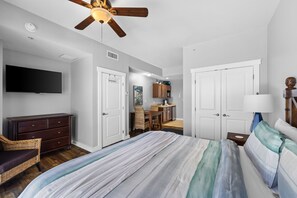 Ein Schlafzimmer mit Bett, Kommode, Fernseher, Deckenventilator und einer kleinen Küche im Hintergrund.