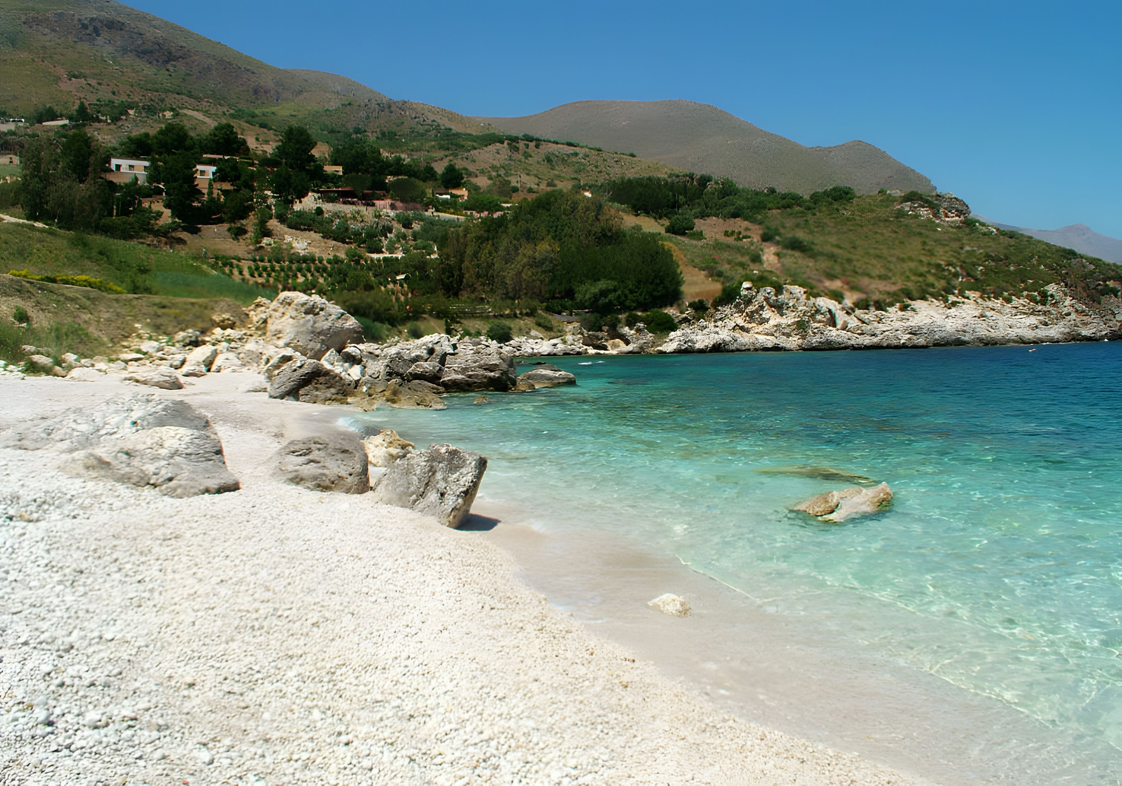 A coastal landscape with a rocky beach, clear turquoise water, and a backdrop of hills.