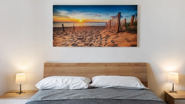 Un dormitorio con cabecera de madera, cama con almohadas blancas y un cuadro colgado en la pared de una playa al atardecer.
