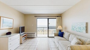 A hotel room with a television, a sofa, a painting, and a view of the sea through a sliding door.