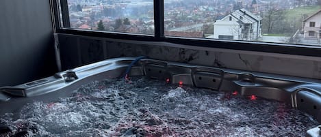 A hot tub with a view of a town and mountains through large windows.