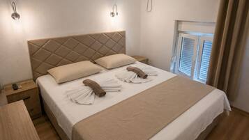 Een opgemaakt bed met hoofdeinde, een nachtkastje met een afstandsbediening en een raam met jaloezieën.