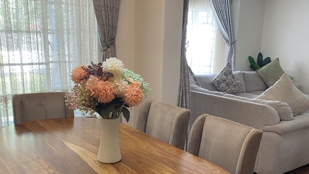 Een eetruimte met een ovale houten tafel, beiges bekleden stoelen en een bloemenvaas.