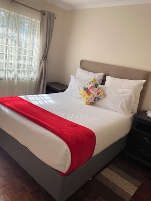 A hotel room with a bed, a nightstand, a red throw blanket, and a window with curtains.