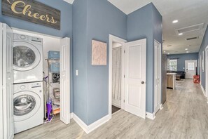 A laundry room with stacked washers and dryers, a door leading to a kitchen, and a hallway with a 'Cheers' sign. - Modern West Tampa Bungalow - Live & Work In Style (Tampa)