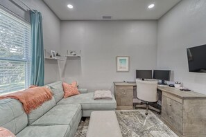 Een moderne woonkamer met een hoekbank, een bureau met twee monitoren, een televisie en een klein plankje met decoratieve spullen.