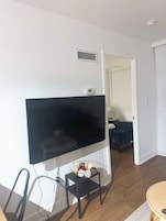 Una sala de estar con un televisor de pantalla plana montado en la pared, una mesita con adornos, y una puerta que da a otra habitación.
